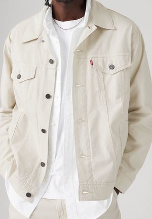 Homme portant une veste beige clair boutonnée par-dessus une chemise blanche et un t-shirt, les mains dans les poches, debout devant un fond neutre.