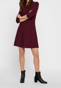 Bordeaux gestricktes Kleid mit langen Ärmeln, A-Linie Silhouette und runder Ausschnitt. Modell kombiniert mit schwarzen Stiefeletten.