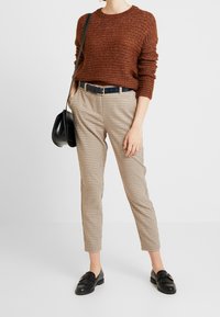 Braun strukturierter Strickpullover, karierte beige- und braunfarbene Hose, schwarzer Gürtel und schwarze Slipper. Modell hält eine schwarze Umhängetasche.