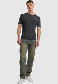 Mørk grå t-skjorte med kort erm og rund hals, med en liten hvit logo på brystet, kombinert med lyse, falmede blå jeans og hvite joggesko.