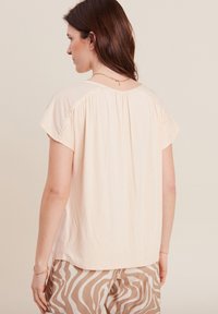 Pfirsichfarbene Bluse mit kurzen Ärmeln und texturierten Häkel-Details auf der Rückseite. Weicher Stoff mit lässigem Fall, kombiniert mit gemusterten Hosen.