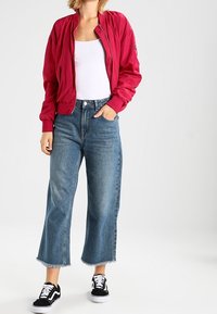 Czerwona kurtka bomberka, biały top na ramiączkach, niebieskie krótkie dżinsy z postrzępionymi końcami, czarne trampki z białymi akcentami. Swobodny strój z warstwowym wyglądem.