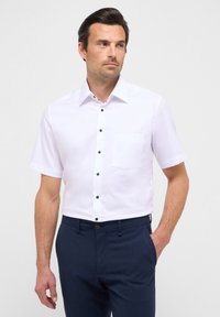 Wit overhemd met korte mouwen, een knoopsluiting aan de voorkant, donkere knopen en een borstzak. Gecombineerd met marineblauwe broek van een gladde stof.