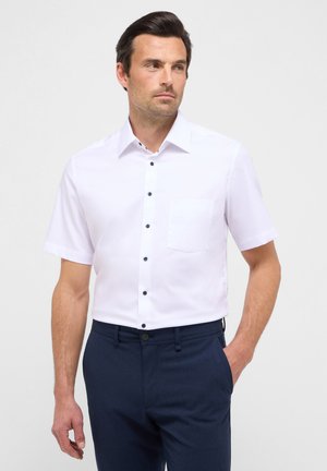 Short-sleeve white shirt with a button-up front, dark buttons, and a chest pocket. Paired with navy blue trousers, smooth fabric texture.