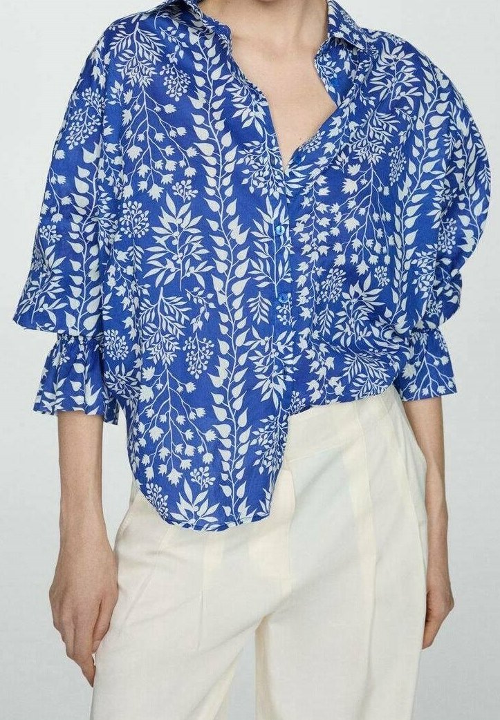 Chemise à boutons bleue à motifs floraux blancs, coupe ample, manches retroussées et poche poitrine. Associée à un pantalon de couleur claire.