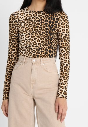 Mujer con una blusa de manga larga con estampado de leopardo metida en unos pantalones beige de cintura alta y pernera ancha, de pie frente a un fondo liso.