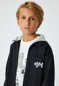 Zwarte denimjack met grijze capuchon, vooraan met rits en een linker borstzak met een wit geborduurd logo. Witte graphic t-shirt eronder.