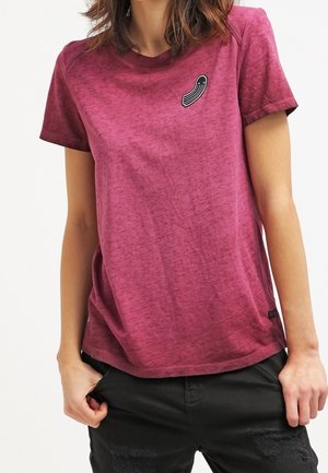 Osoba ubrana w bordo koszulkę z krótkim rękawem z małą czarno-białą naszywką na lewej piersi, w połączeniu z czarnymi spodniami.