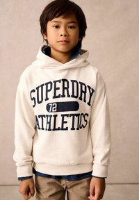 Krémová mikina s kapucí s potiskem "SUPERDRY ATHLETICS" v námořnické modři. Model má pod ní na sobě džínovou košili. Jsou vidět zapnuté béžové kalhoty. Měkká textura.