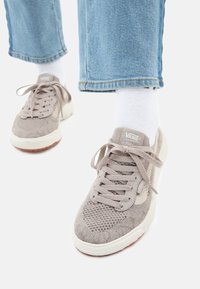 Beige Vans sportcipők kötött textúrával, fűzős dizájnnal, fehér kiegészítőkkel és gumi talppal. Világoskék farmerral és fehér zoknival viselve.