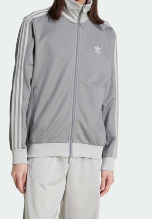 Persona che indossa una giacca sportiva Adidas grigio chiaro con zip e tre strisce bianche sulle maniche, abbinata a pantaloni grigio chiaro.