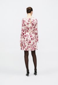 Γυναίκα με μαλλιά σε κότσο, ντυμένη με λευκό φόρεμα με μακριά μανίκια και άνθη, μαύρο καλσόν και μαύρες γόβες, στέκεται γυρισμένη ανάποδα σε λευκό φόντο.