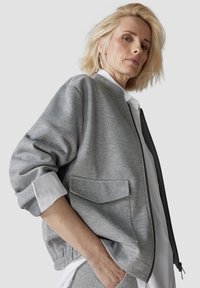 Gray bomber jacket made of textured fabric, featuring a front zipper, ribbed cuffs, and two patch pockets. Styled over a white shirt.