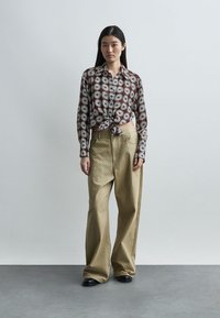 Γυναίκα με καφέ πουκάμισο με λουλούδια, κουμπωτό και δεμένο, φόρεμα με μπεζ παντελόνι φαρδύ και μαύρα παπούτσια, στέκεται μπροστά σε μονόχρωμο ανοιχτό γκρι φόντο.