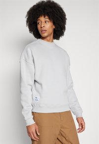 Young man with curly hair wearing a light grey sweatshirt and brown pants, looking sideways against a plain background.