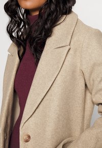 Abrigo de mezcla de lana beige con solapas de muesca, cierre de un solo botón y tejido texturizado. Llevado sobre una blusa de canalé burdeos.