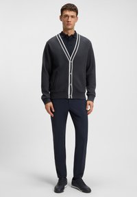 Tummanharmaa neuletakki valkoisilla reunoilla, napillinen etuosa, sivutaskut, yhdistetty tummansinisiin housuihin ja tummiin kenkiin. Sileä pinta, rento malli.