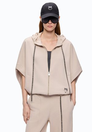 Mujer vistiendo sudadera con capucha beige con cremallera y cordones de cadena negros, pantalones a juego con detalle de cadena negra, gorra negra y grandes gafas de sol negras.