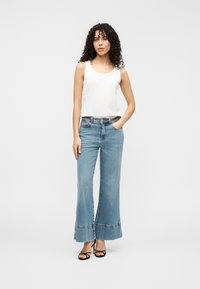 Λευκό αμάνικο τοπ συνδυασμένο με ανοιχτόχρωμα μπλε τζιν με φαρδιά παντελόνια, που διαθέτουν σχέδιο στο τελείωμα και σκούρα ζώνη. Φορεμένο με μαύρα πέδιλα με λουράκια.