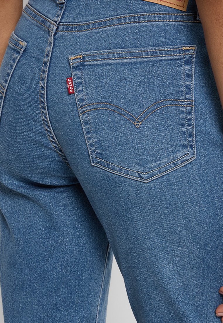 Jean en denim bleu avec une poche arrière visible présentant un motif de couture courbé distinctif et un petit logo rouge sur la poche droite.