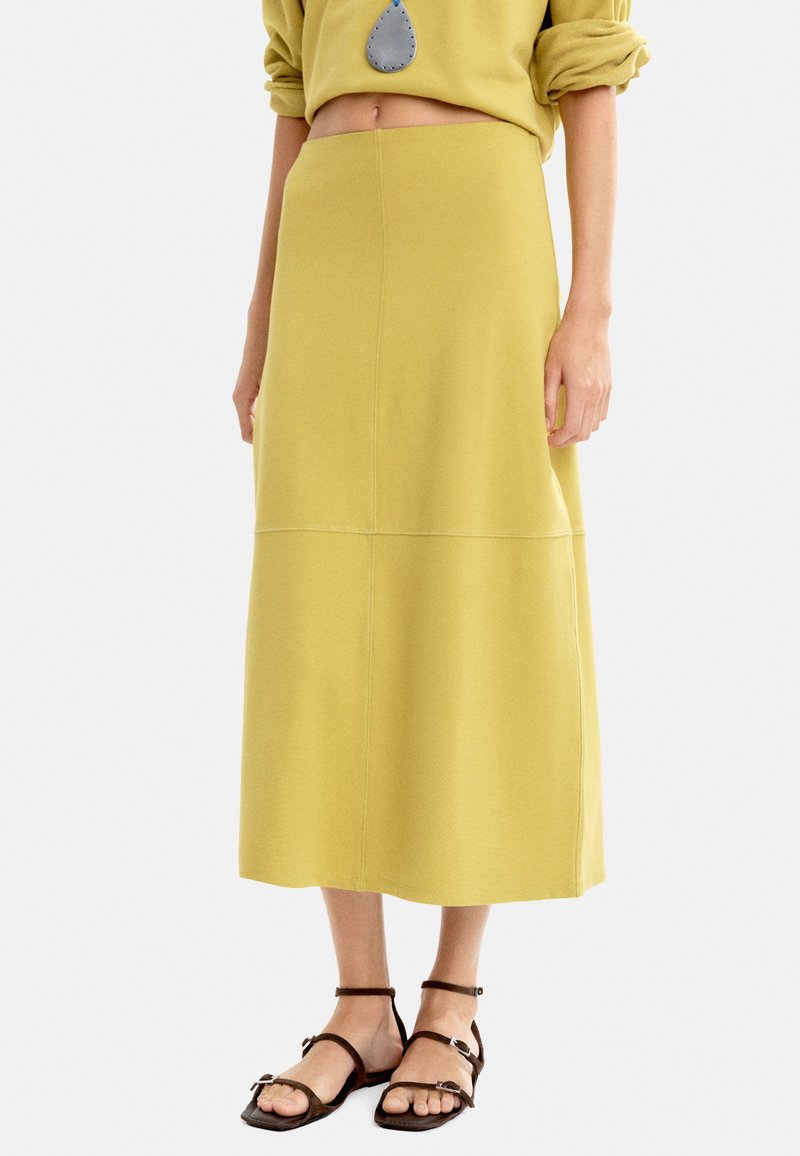 Gelbe Midi-Skirt aus weichem Material, mit geradem Schnitt und Nahtdetails, kombiniert mit braunen Riemchensandalen.