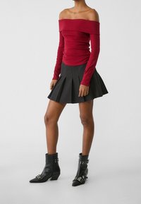 Haut rouge côtelé à manches longues et épaules dénudées, associé à une mini-jupe noire plissée et des bottines noires à bouts pointus avec boucles décoratives.