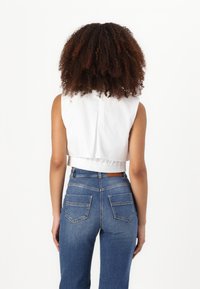 Top corto bianco senza maniche con fondo arricciato, abbinato a jeans in denim blu a vita alta con tasche posteriori e un'etichetta marrone.