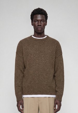 Young Black man facing forward wearing a brown speckled knit sweater over a white shirt and beige pants against a plain light background.