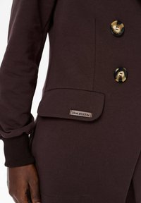 Bruine sweatshirt met twee grote knoopsluitingen, een patchzak en een "blue shadow" label. De stof lijkt zacht en gestructureerd.