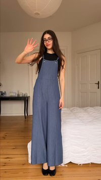 Νέα γυναίκα με μακριά σκούρα μαλλιά και γυαλιά, φορά μπλε φαρδιά σαλοπέτα και χαιρετά ενώ στέκεται σε μια κρεβατοκάμαρα με ξύλινο δάπεδο.