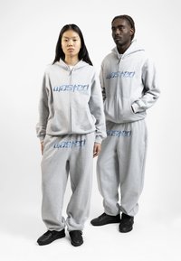 Grå hættetrøjer og bukser har blå "wasted" tekst. Stoffet ser blødt ud, med en afslappet pasform og sidelommer, kombineret med sorte sneakers.