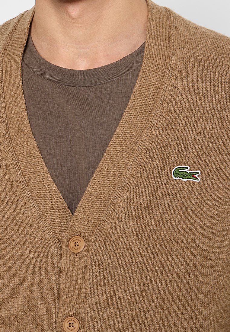 Cardigan en tricot marron avec un col en V, doté de boutons en bois et d'un petit logo de crocodile vert sur le côté gauche.