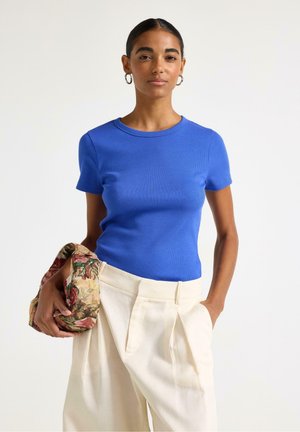 Kvinde iført en blå kortærmet top og løse cremefarvede bukser, der holder en clutchtaske med blomstermønster i den ene hånd.