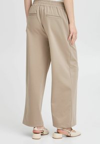 Pantalons larges en tissu taupe clair, dotés d'une taille élastique, de deux poches arrière et d'une texture lisse. Associés à des sandales à talons blocs beiges.