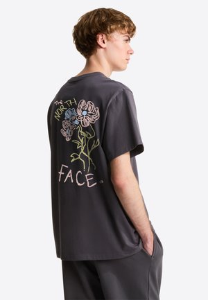 Jonge man draagt een donkergrijs T-shirt met een bloemmotief en de tekst "The North Face" op de rug, staand met handen in de zakken.