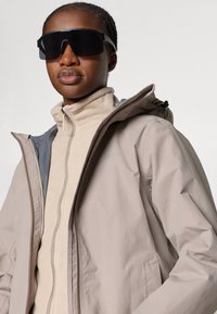 Beige zips-šaty s vysokým golierom, sivou vnútornou podšívkou a kapucňou. Čierne oversized slnečné okuliare. Hladká textúra látky.