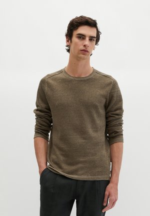 Jeune homme aux cheveux bouclés foncés portant un pull en maille vert olive à manches longues et un pantalon noir, debout les mains dans les poches sur fond blanc.