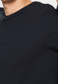 Granatowa bawełniana koszulka z okrągłym dekoltem. Gładka tekstura i krótkie rękawy, odsłaniające część dżinsowej kurtki na lewym ramieniu.