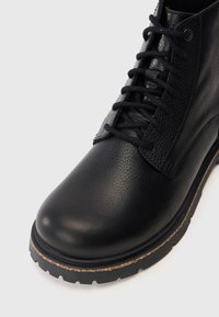 Stivale in pelle nera con finitura testurizzata, punta tonda e suola in gomma marrone a contrasto. Presenta un design con lacci e dettagli sui pannelli laterali.