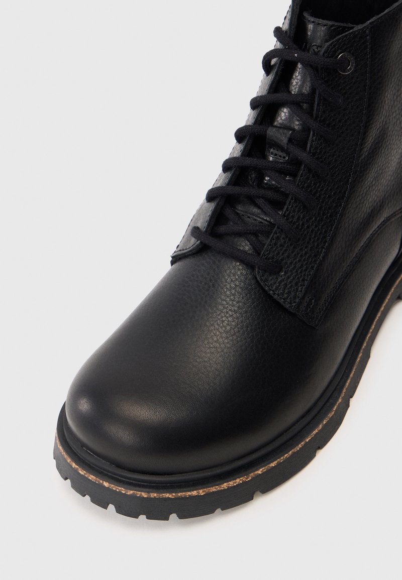 Sort læderstøvle med struktureret finish, rund tå og kontrasterende brun gummisål. Har snørebåndsdesign og detaljer i sidepanelet.