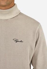 Beżowy sweter z kołnierzem typu golf, z prążkowanym wzorem, z wysokim kołnierzem i małym czarnym haftowanym logo na lewej części klatki piersiowej. Miękka tekstura.