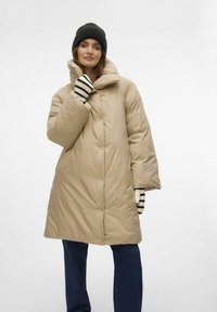 Beige dunjakke med høj krave, knaplukninger og oversized pasform. Båret med en sort hue og stribede handsker mod en hvid baggrund.