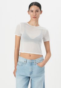 Prozirni bijeli kratki top s kratkim rukavima, s okruglim izrezom i laganim slojevima preko sive bralette, u kombinaciji s svijetloplavim farmerkama.