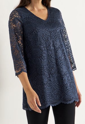 Kobieta nosząca granatową koronkową tunikę z rękawami do łokcia i falowanymi brzegami na czarnych spodniach, widoczna od szyi do bioder.