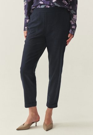 Donna che indossa pantaloni scuri blu navy a gamba stretta con orli risvoltati, abbinati a décolleté beige con cinturino slingback e una maglia a maniche lunghe viola con fantasia floreale.