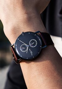Montre analogique noire avec deux sous-cadrans et accents argentés, montée sur un bracelet en cuir marron foncé. Le cadran présente des marqueurs d'heure clairs.