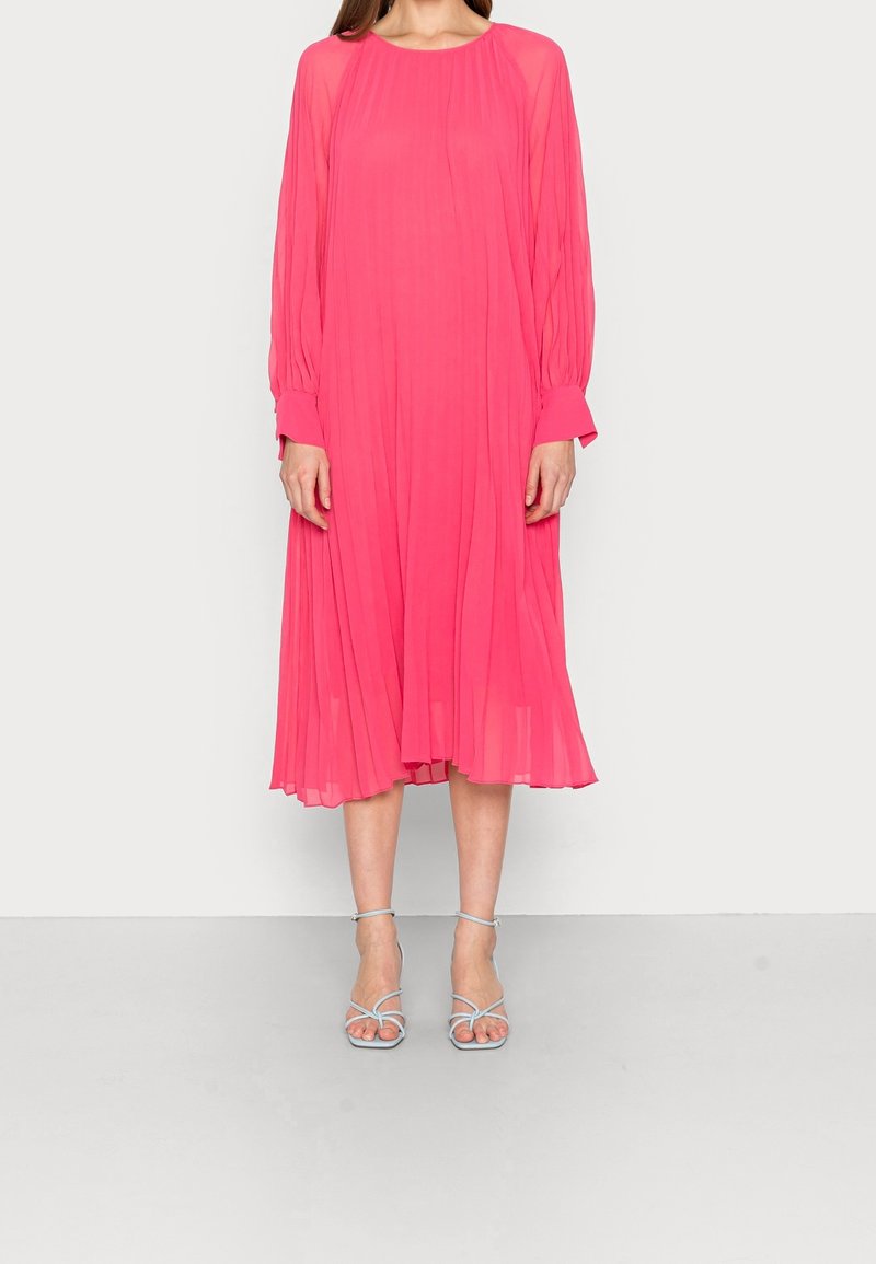 Femme portant une robe midi rose vif à manches longues et des sandales à talons hauts à brides bleu clair, debout devant un fond uni.