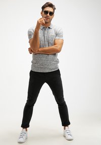 Grijze marl polo met korte mouwen, gecombineerd met zwarte slim-fit pantalon en witte sneakers met drie strapsluiting. Zwarte zonnebril.
