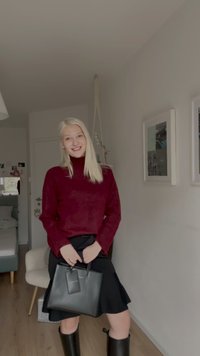 Svart läderhandväska med en tagg, bärs med en vinröd stickad tröja och svart kjol, ihop med långa svarta gummistövlar. Ljust inredd rum.
