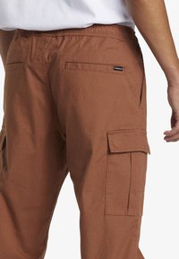 Hnědé cargo kalhoty s elastickým pasem, texturovaná tkanina, boční kapsy a kapsa na stehně se klopou. Viditelná decentní značka.